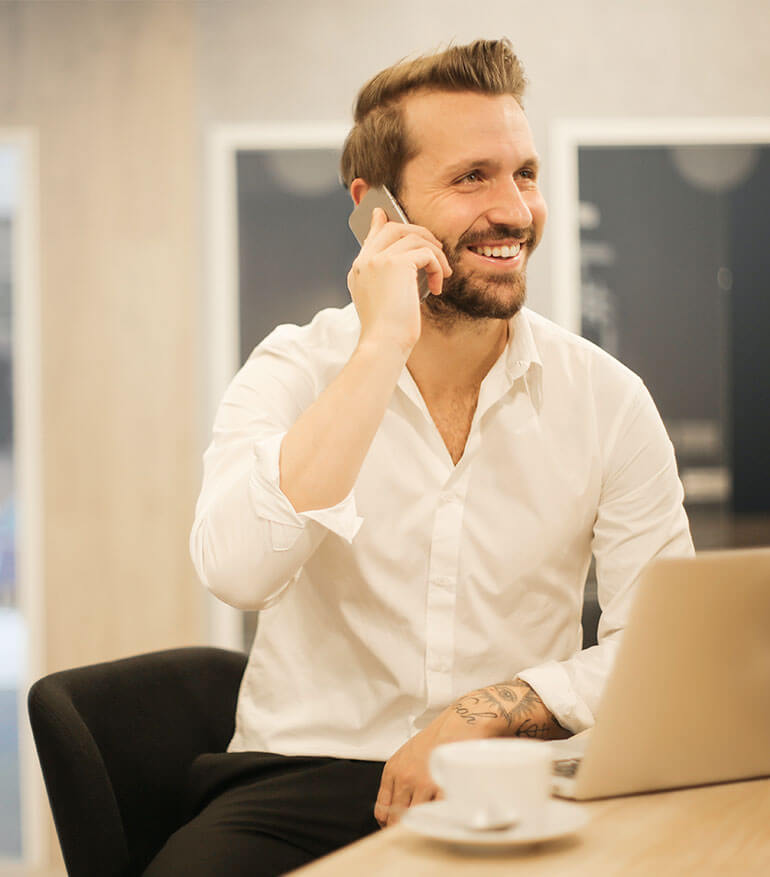 Man in business clothes smiling on a cell phone, sitting in front of a laptop